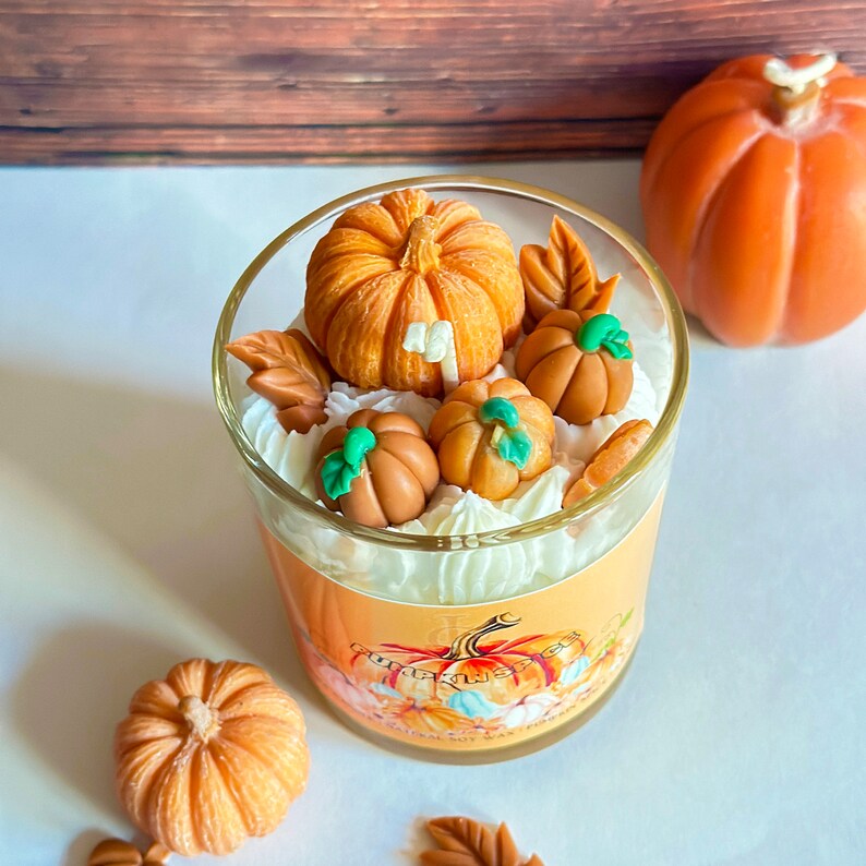 May include: A glass jar candle with a white and orange pumpkin design. The candle is topped with white frosting and decorated with small orange and green pumpkin figurines.
