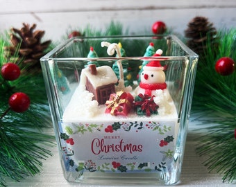 Pode incluir: Um castiçal quadrado de vidro com uma cena de Natal. No interior, um boneco de neve, uma casa e árvores assentam sobre uma base nevada. A vela tem um rótulo que diz "Feliz Natal". Bagas vermelhas e ramos de pinheiro rodeiam a vela.