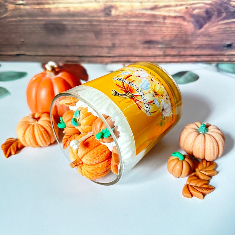 May include: A glass jar candle with a yellow label that says "Pumpkin Spice" and a white background. The label has a drawing of a pumpkin with green leaves. The candle is surrounded by small orange and brown pumpkins.