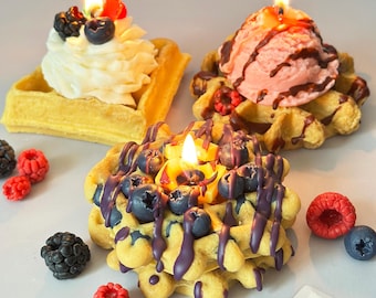 May include: Three waffle-shaped candles with lit flames. One candle is topped with whipped cream and berries, another with pink ice cream and chocolate drizzle, and the third with blueberries and purple drizzle. Scattered around are berry-shaped decorations.