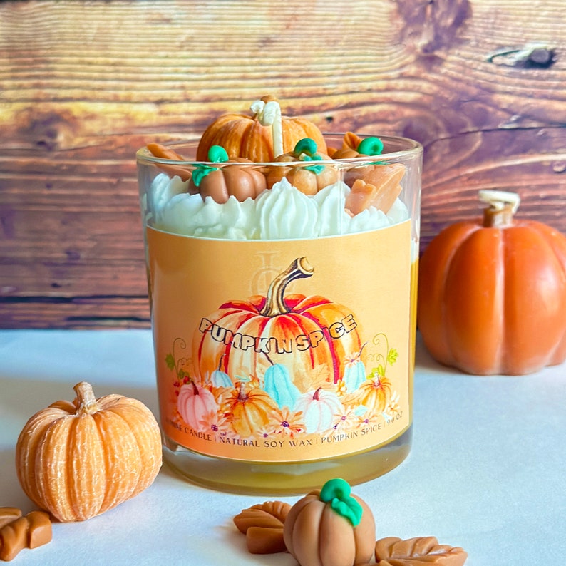 May include: A glass candle jar with a white lid and a label that says "Pumpkin Spice" with a pumpkin illustration. The candle is surrounded by small decorative pumpkins and leaves.