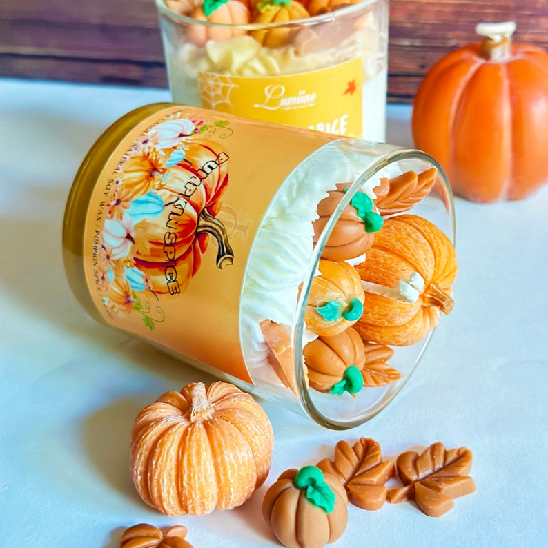 May include: A glass jar candle with a white lid and a label that says "Pumpkin Spice" with a pumpkin graphic. The candle is surrounded by small orange pumpkin-shaped decorations and brown leaf decorations.