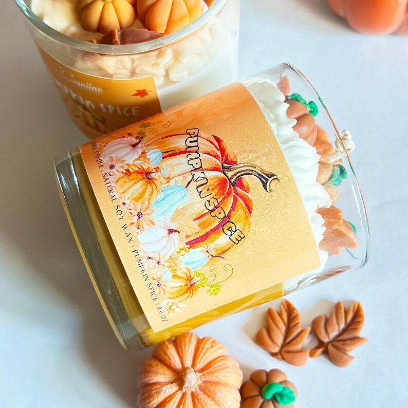 May include: A yellow candle with a white lid and a pumpkin spice label. The candle is surrounded by small orange pumpkins and brown leaves. The label says "Pumpkin Spice" and "100% Natural Soy Wax - Pumpkin Spice 9 oz."