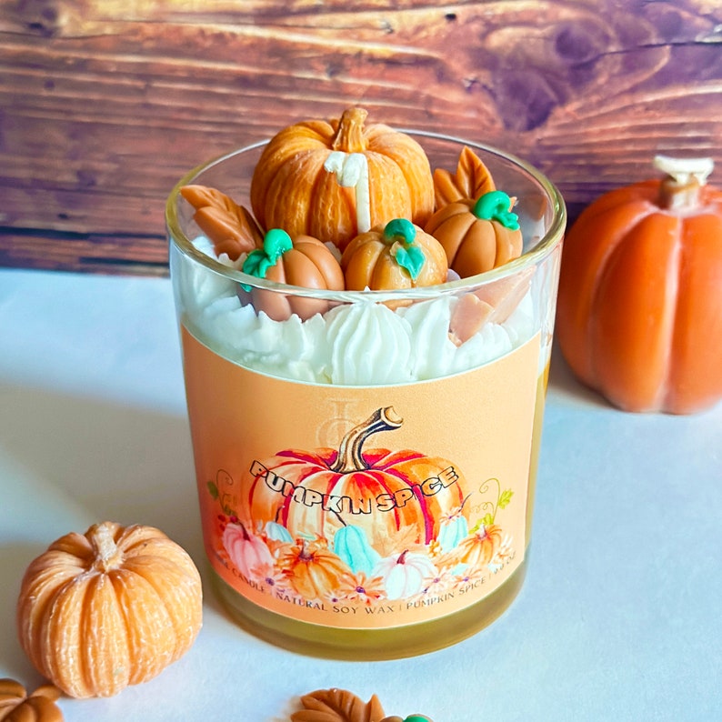 May include: A glass jar candle with a pumpkin spice scent. The candle is topped with a white frosting and decorated with small orange and green pumpkin-shaped candies. The label on the jar reads "Pumpkin Spice" and features a watercolor illustration of pumpkins and leaves.