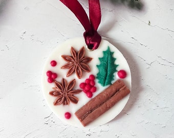 May include: A circular, white Christmas decoration with a burgundy ribbon. It features brown star anise, red berries, a green holly leaf, and brown cinnamon sticks. A festive holiday ornament.