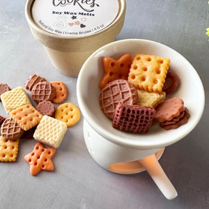 Könnte beinhalten: Ein weißer Keramik-Wachsschmelzer mit einem kleinen Griff. Der Schmelzer ist mit verschiedenen keksförmigen Sojawachs-Schmelzen in Braun, Orange und Gelb gefüllt. Die Schmelzen sind in einer schalenförmigen Schale auf dem Schmelzer angeordnet. Der Schmelzer ist mit "Soy Wax Melts" und "Creme Brulee 1.4 oz." beschriftet.