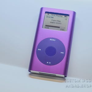 May include: A purple iPod digital media player showing "Now Playing" on its screen, with song information. It features a circular control pad with a central button and navigation controls. The text "CUSTOM IPODS ANDRD.SHOP" is visible.