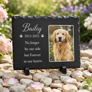 May include: A rectangular black slate memorial plaque featuring a photo of a golden retriever. The text reads: Bailey, 2013-2025, No longer by our side but Forever in our hearts, with two paw print icons. The plaque is supported by two black stands.