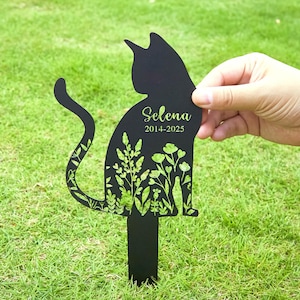 May include: Black metal cat-shaped garden stake with the name "Selena" and dates "2014-2025" in green script. The cat silhouette features a floral design in green. The stake is held in a grassy area.
