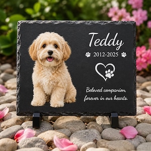 May include: A rectangular black slate memorial plaque featuring a printed image of a light-colored fluffy dog. The text reads Teddy, 2012-2025, and Beloved companion, forever in our hearts, alongside a heart icon with a small paw print inside.