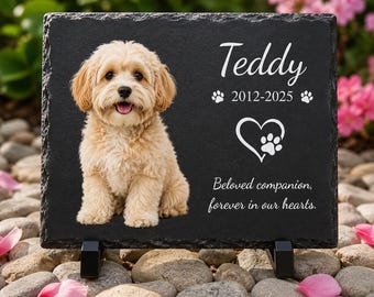 May include: A rectangular black slate memorial plaque featuring a printed image of a light-colored fluffy dog. The text reads Teddy, 2012-2025, and Beloved companion, forever in our hearts, alongside a heart icon with a small paw print inside.