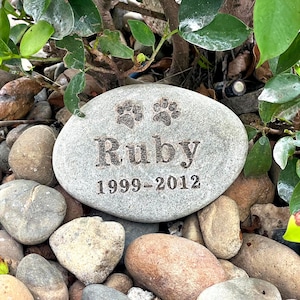 May include: A gray, oval-shaped memorial stone engraved with the name "Ruby", paw prints, and the years "1999-2012". The stone is surrounded by pebbles and greenery, creating a natural setting.