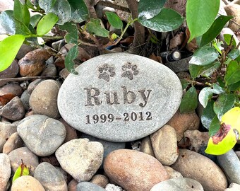 May include: A gray, oval-shaped memorial stone engraved with the name "Ruby", paw prints, and the years "1999-2012". The stone is surrounded by pebbles and greenery, creating a natural setting.