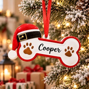 May include: A bone-shaped Christmas ornament with a white centre, red border, and a red Santa hat with a black belt on the left side. The name Cooper is printed in black script in the centre, flanked by two brown paw prints. It hangs by a red ribbon.