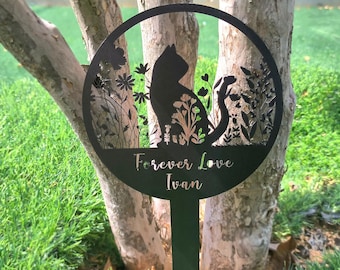 May include: Black metal garden stake featuring a cat silhouette within a circular frame, surrounded by floral and plant cutouts. The text "Forever Love Ivan" is inscribed below the cat. The stake is placed in a grassy area near a tree.