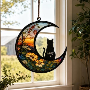 May include: A crescent moon-shaped stained glass suncatcher featuring a black silhouette of a cat sitting on the moon. The cat has the name Lucy and a small heart printed on its body. The glass depicts a sunset landscape with orange flowers.