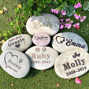 May include: A selection of engraved memorial stones for pets, each featuring a name, paw print, and dates. The stones vary in size and shape, with names such as "Casey," "Emma," and "Molly" etched in different fonts.