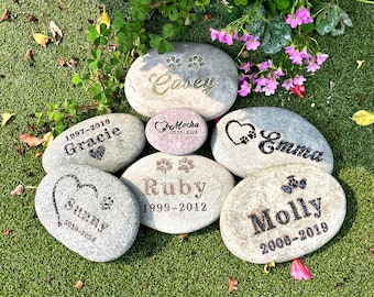 May include: A collection of engraved memorial stones for pets, each with a name, paw print, and dates. The stones are various sizes and shapes, with names like "Casey," "Emma," and "Molly" etched in different fonts.
