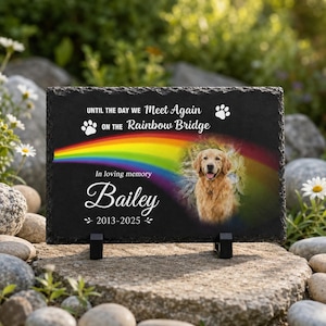 May include: A rectangular black slate memorial plaque featuring a printed rainbow, a golden retriever portrait, and the text: UNTIL THE DAY WE MEET AGAIN ON THE RAINBOW BRIDGE, In loving memory, Bailey, 2013-2025, with two white paw prints.