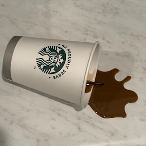 May include: A white paper coffee cup with a gray rim and the Starbucks logo is tipped over, spilling brown coffee onto a light-colored surface. A black straw is visible in the cup and the spilled coffee.