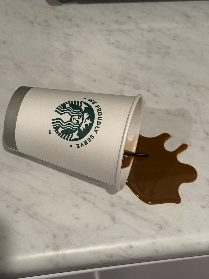 May include: A white paper coffee cup with a gray rim and the Starbucks logo is tipped over, spilling brown coffee onto a light-colored surface. A black straw is visible in the cup and the spilled coffee.