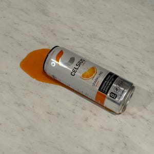Może przedstawiać: Srebrna puszka napoju energetycznego Celsius Sparkling Orange, częściowo rozlana, z pomarańczowym płynem na jasnej powierzchni. Puszka zawiera logo Celsius i grafikę plasterka pomarańczy.