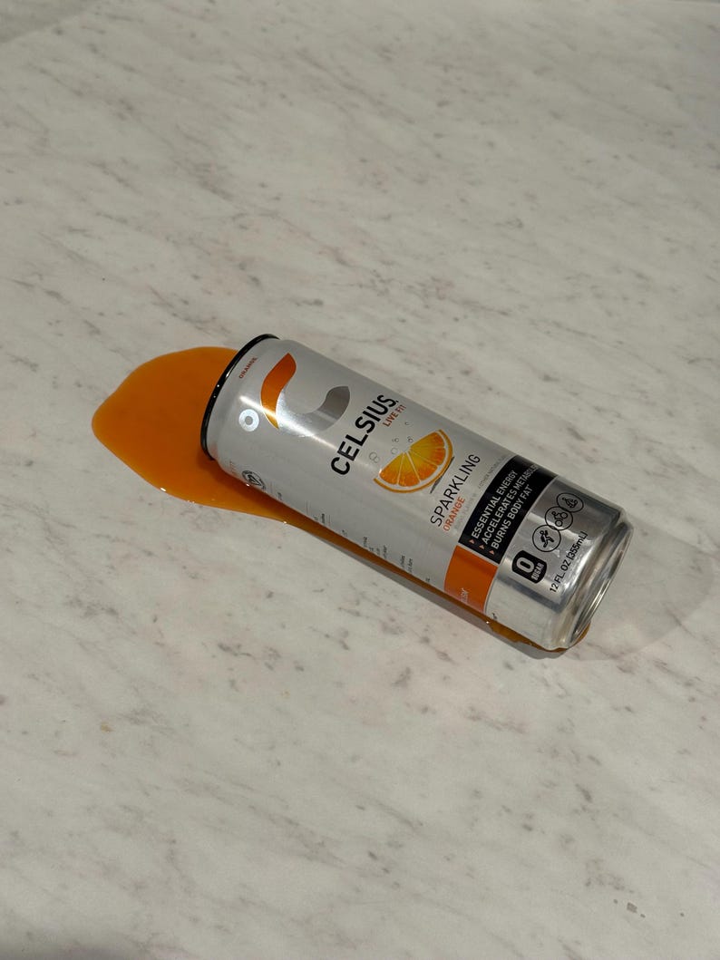 Może przedstawiać: Srebrna puszka napoju energetycznego Celsius Sparkling Orange, częściowo rozlana, z pomarańczowym płynem na jasnej powierzchni. Puszka zawiera logo Celsius i grafikę plasterka pomarańczy.