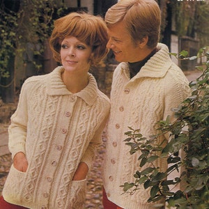 May include: Two people wearing cream-colored cable knit cardigans with button closures. The cardigan has two pockets on the front. The woman is wearing red pants and the man is wearing red pants. The background is a blurry image of a brick wall and green plants.