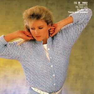 May include: A light blue knitted cardigan with a button-up front and a white belt. The cardigan has a lacy pattern and is shown on a model with blonde hair. The text "Patons promise DOUBLE KNITTING" is at the top of the image. The text "PRICE CODE B 7992 CANADA $1.00 76-102 cm 30-40 in Teens sizes included" is at the bottom of the image.