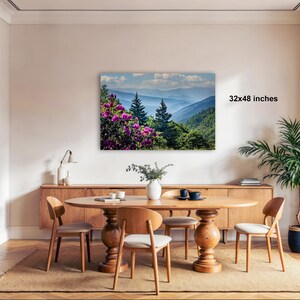 a dining room with a large wooden table surrounded by chairs, a vase of flowers, and a painting of a mountainous landscape with pink flowers hanging on the wall.