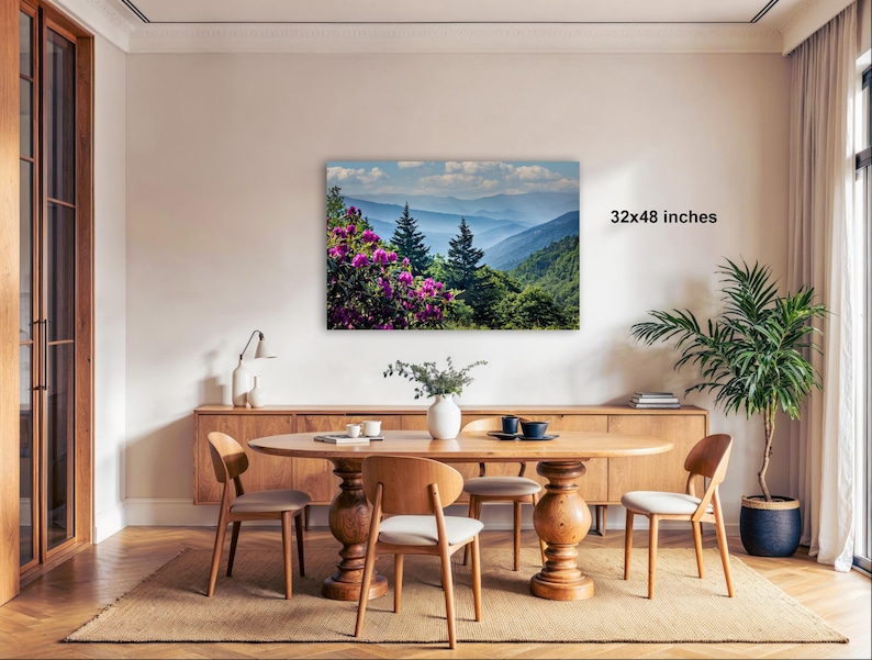 a dining room with a large wooden table surrounded by chairs, a vase of flowers, and a painting of a mountainous landscape with pink flowers hanging on the wall.