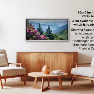 a living room with a wooden coffee table, two white armchairs, and a potted plant. A framed landscape photograph is hanging on the wall, and there is text providing information about the frame options.