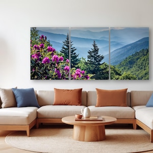 a living room with a large, colorful painting of a mountain landscape hanging on the wall. The painting is composed of three panels, depicting a serene mountain scene with trees, flowers, and a clear blue sky. The room is furnished with a beige sofa and a wooden coffee table, creating a cozy and inviting atmosphere.
