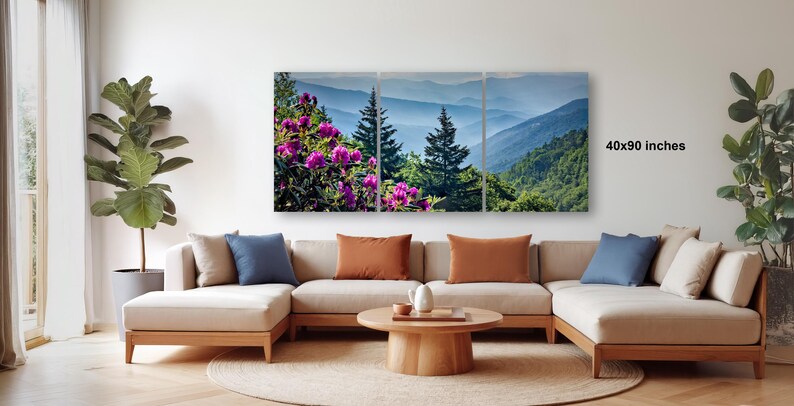 a living room with a large, colorful painting of a mountain landscape hanging on the wall. The painting is composed of three panels, depicting a serene mountain scene with trees, flowers, and a clear blue sky. The room is furnished with a beige sofa and a wooden coffee table, creating a cozy and inviting atmosphere.