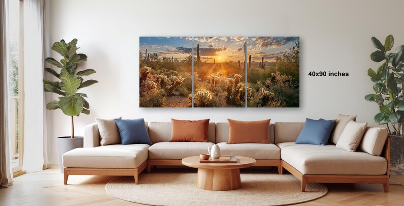 a living room with a large, colorful painting of a desert landscape hanging on the wall. The painting depicts a desert scene with cacti, shrubs, and a sunset. The room has a beige sofa and a wooden coffee table, and there are several potted plants placed around the space.