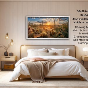 The image is a modern bedroom with a large bed, wooden furniture, and a framed landscape painting hanging on the wall. The painting depicts a desert scene with a sunset. The room has a minimalist and clean design, with a white color scheme and natural elements like a vase and a plant.
