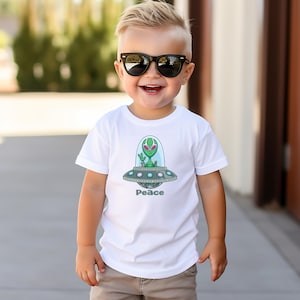 May include: A white t-shirt featuring a cartoon alien in a spaceship with the word "Peace" below. The alien is green with a large head and is waving. The spaceship is gray and blue. The t-shirt is worn by a young child wearing sunglasses.