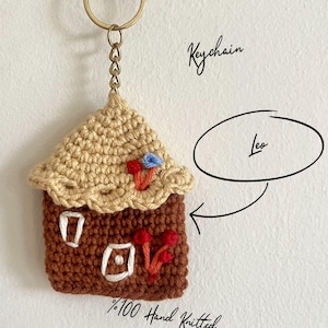 May include: A handmade, brown and beige knitted house keychain with a gold-colored keyring. The house features white window details, red and blue floral accents, and the text "Leo". The keychain is labeled as "100% Hand Knitted".