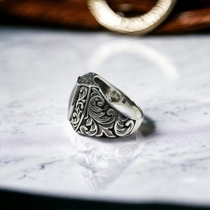 May include: A silver ring with an ornate design featuring a dark gray stone. The ring is set on a white marble surface.
