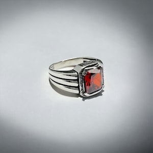 May include: A silver ring with a red gemstone set in a square bezel. The ring has a textured band with three raised lines.