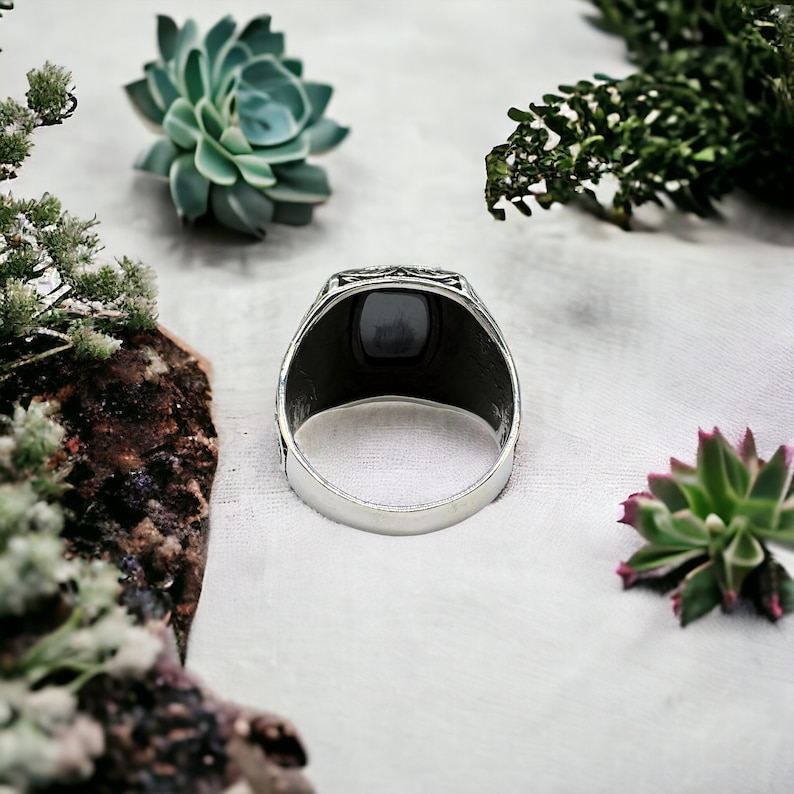 May include: A silver ring with a black stone set in a decorative bezel. The ring is on a white background.