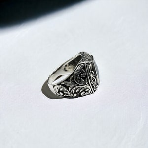 May include: A silver ring with an ornate design and a dark gray stone in the center.
