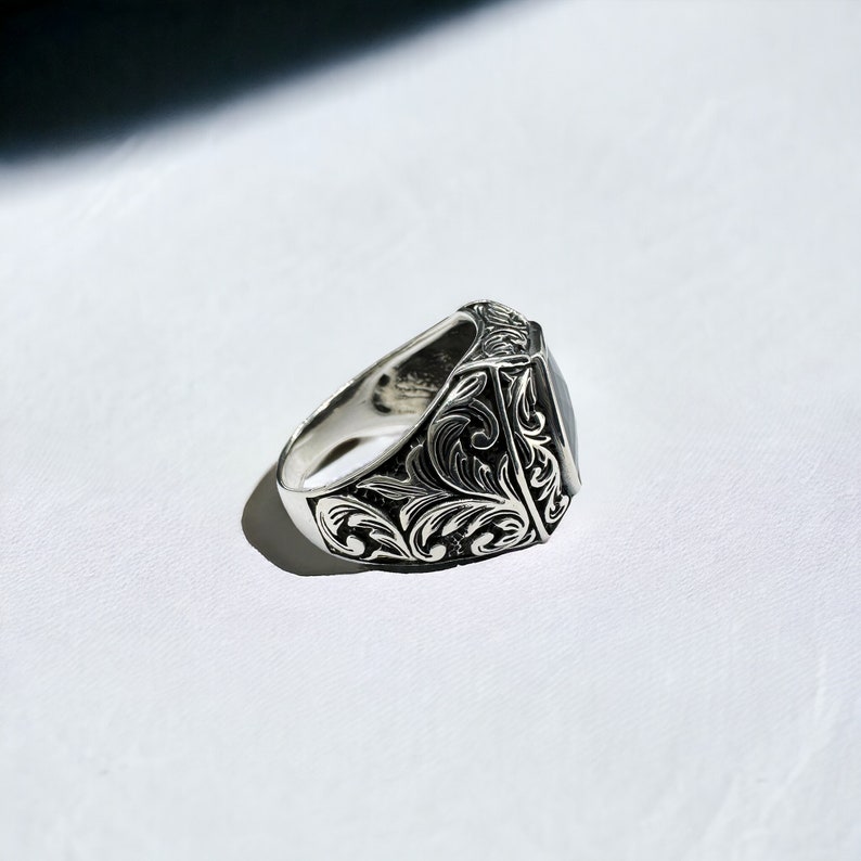 May include: A silver ring with an ornate design and a dark gray stone in the center.