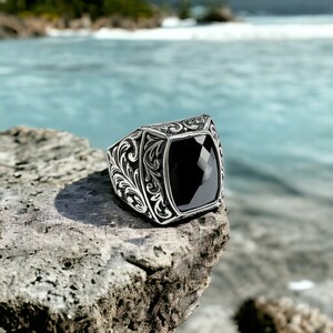 May include: A silver ring with an ornate design and a black gemstone. The ring is sitting on a rock with a blurry background of water.