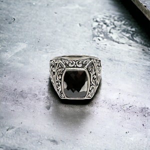 May include: A silver ring with a black gemstone set in a square shape. The ring has an intricate floral design around the gemstone.