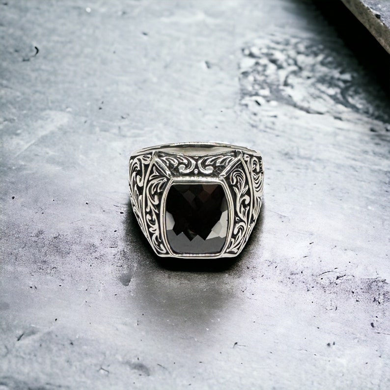 May include: A silver ring with a black gemstone set in a square shape. The ring has an intricate floral design around the gemstone.
