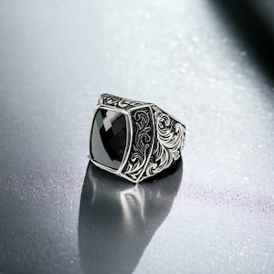 May include: A silver ring with an ornate design featuring a black gemstone. The ring has a square-shaped setting with a detailed floral pattern.