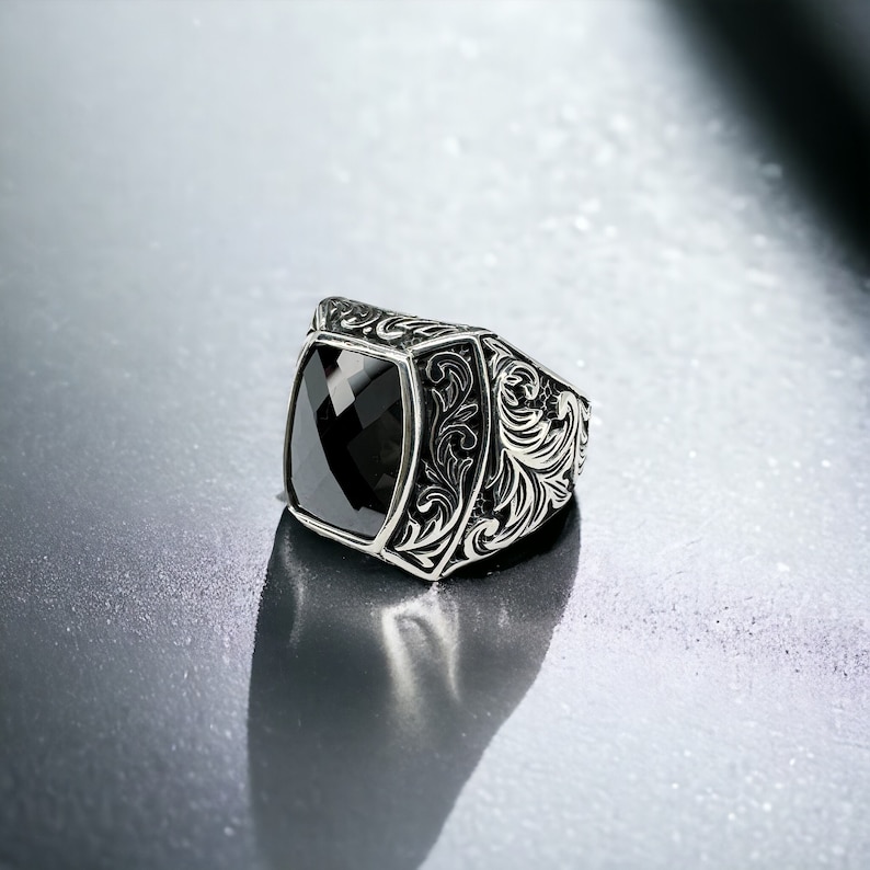May include: A silver ring with an ornate design featuring a black gemstone. The ring has a square-shaped setting with a detailed floral pattern.