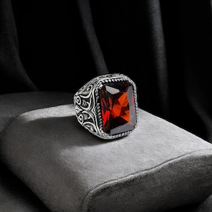 May include: A silver ring with an ornate design and a large, square, red gemstone.