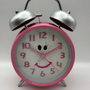 Op de afbeelding: Roze wekker met een lachend gezichtsontwerp. De klok heeft een zilveren bel en handvat, met roze cijfers en wijzers op een witte wijzerplaat. Klassiek ontwerp.